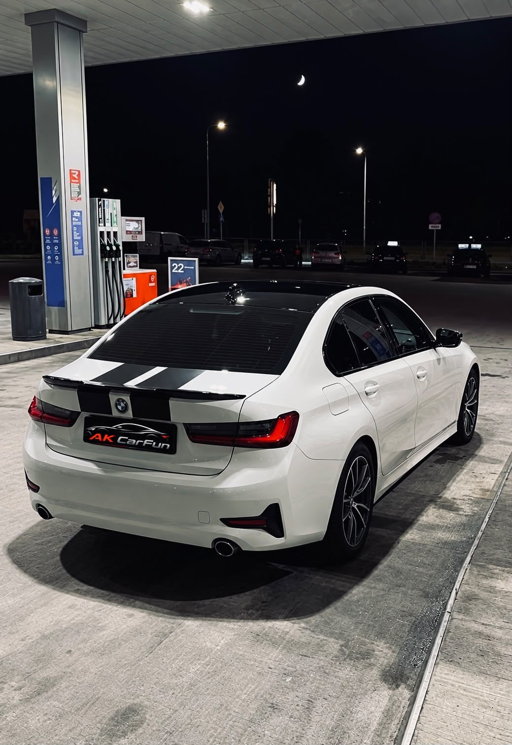 Białe BMW serii 3 z floty AK CarFun nocą na stacji paliw, widok z tyłu na tablicę z logo AK CarFun
